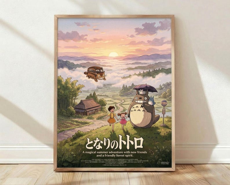 May include: Framed poster print displayed against white wall on wooden floor. Artwork shows beloved forest spirit Totoro with umbrella standing beside two children at rural bus stop, with flying catbus overhead. Mountain landscape background features sunset sky in pink and orange hues with rolling hills and traditional Japanese house. Natural wood frame, soft natural lighting with gentle shadows.