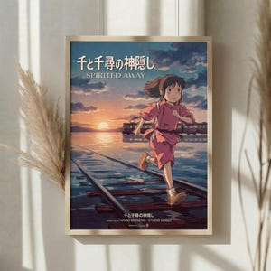 May include: Framed Studio Ghibli Spirited Away movie poster featuring young Chihiro running along flooded train tracks at sunset. The anime-style artwork shows warm orange and blue sky colors reflecting in water, with a distant train. Golden wooden frame hangs on white wall near window with natural light and dried pampas grass decoration visible to the left.