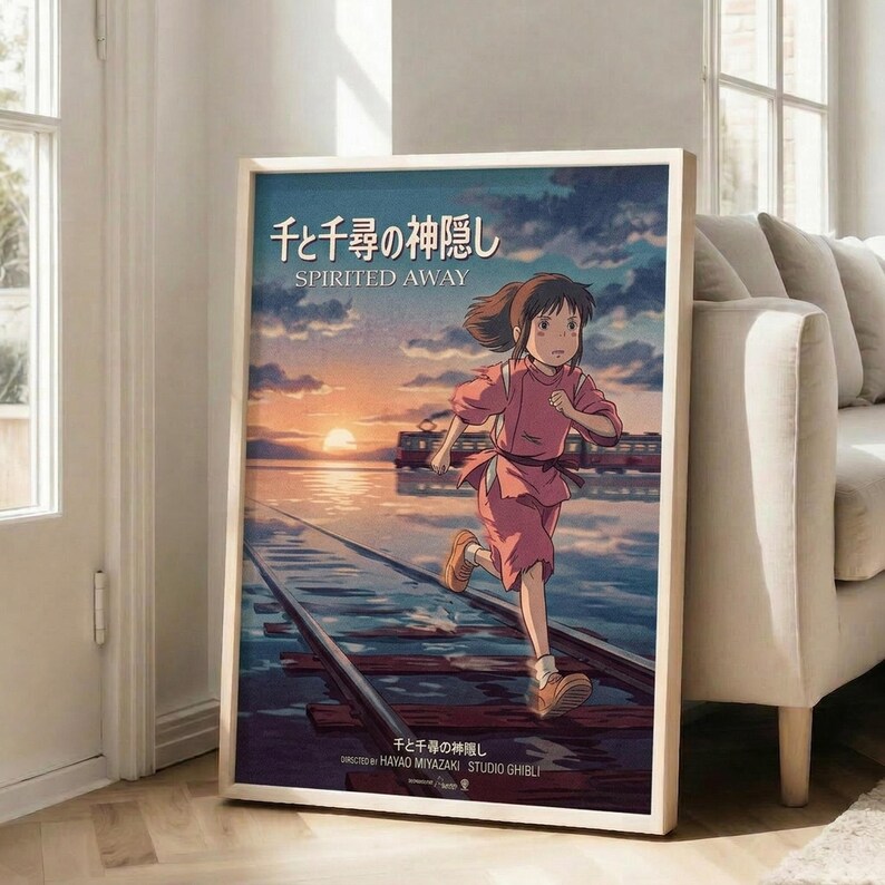 May include: Framed Studio Ghibli Spirited Away movie poster leaning against white wall in bright living room. Shows anime girl with brown hair in pink outfit running on wooden railway tracks over water at sunset. Japanese and English text visible. Beige sofa and windows with natural light in background.