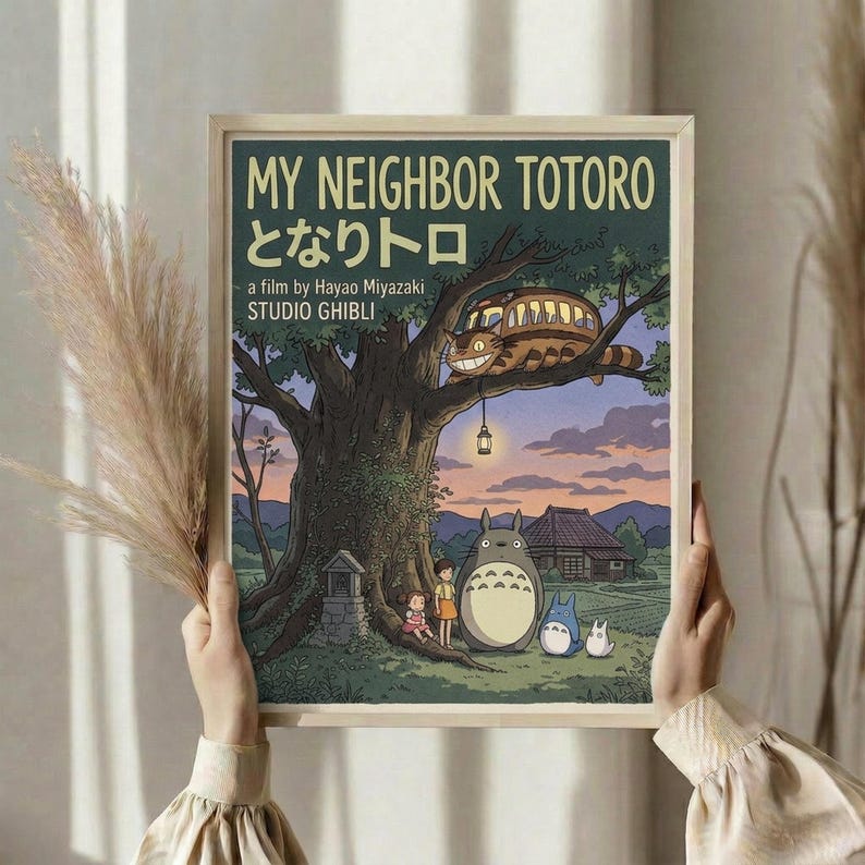 May include: Hands holding a framed poster featuring Totoro characters under a large tree with the iconic cat bus perched in branches above. The artwork shows a twilight scene with mountains, traditional buildings, and small human figures. Dried wheat stalks visible on the left side. The poster has cream-colored matting in a light wood frame against white curtains.