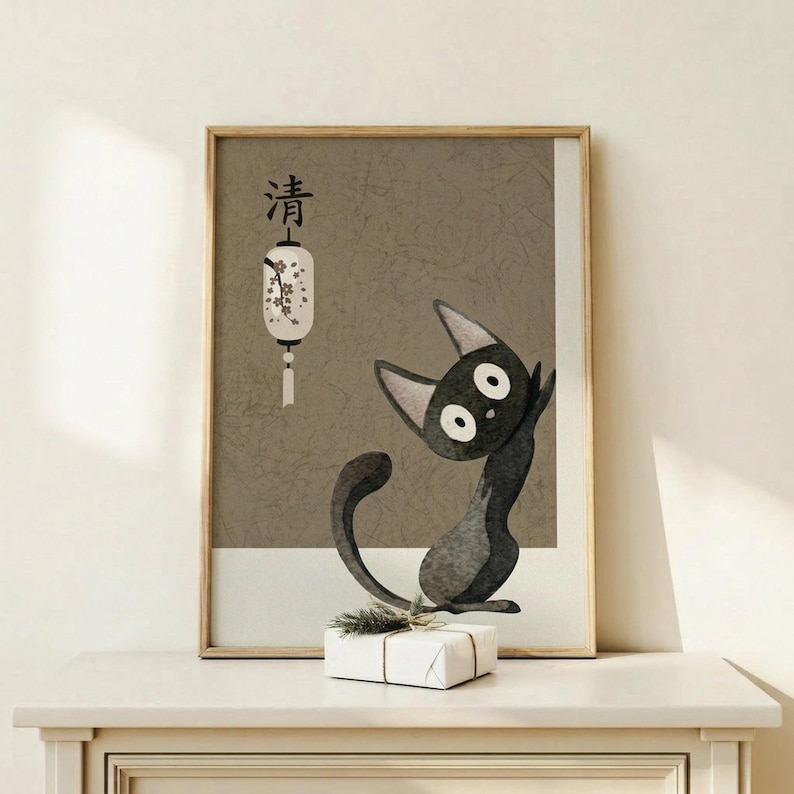 May include: Framed poster print featuring black cat character with large white eyes and pointed ears in Japanese art style, sitting beside white paper lantern with floral design and Japanese characters above. Gray textured background with crackled effect. Natural wood frame displayed on light wooden surface with small wrapped gift box decorated with evergreen sprig. Neutral beige wall backdrop.