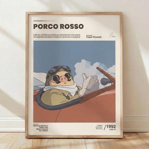 May include: A framed movie poster for Porco Rosso featuring a pig pilot in a red airplane against a blue sky with clouds. The poster includes the title, director Hayao Miyazaki, cast names, the year 1992, and a brief plot summary in black text.