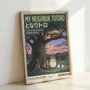 May include: Framed vintage-style movie poster featuring beloved animated characters under a large camphor tree at dusk. The artwork shows forest spirits including a large gray creature and smaller blue companion alongside two children near a traditional Japanese house. A whimsical cat-shaped bus with glowing eyes perches in the tree branches above. The poster has cream text on a dark green background with warm sunset colors throughout the pastoral scene. Light wooden frame displayed against whi