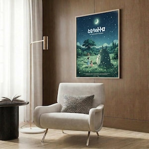 May include: Framed Studio Ghibli My Neighbor Totoro poster mounted on wood-paneled wall above beige armchair with geometric pillow. Poster shows nighttime scene with large gray Totoro character, two children, and crescent moon against starry blue sky over green landscape. Modern reading nook with brass floor lamp and side table.
