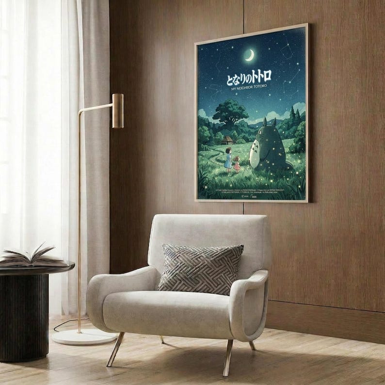 May include: Framed Studio Ghibli My Neighbor Totoro poster mounted on wood-paneled wall above beige armchair with geometric pillow. Poster shows nighttime scene with large gray Totoro character, two children, and crescent moon against starry blue sky over green landscape. Modern reading nook with brass floor lamp and side table.
