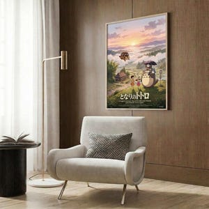 May include: Framed Studio Ghibli Totoro movie poster mounted on wood-paneled wall above beige upholstered armchair with geometric pillow. Poster shows whimsical landscape scene with large gray forest spirit character, children, rural houses, and airship against sunset sky with Japanese text. Modern reading nook includes brass floor lamp and side table with open book.