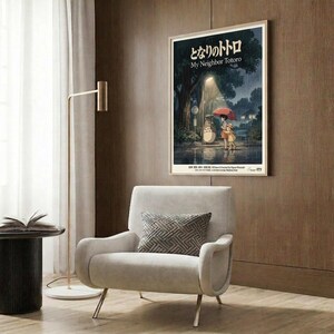 May include: Framed My Neighbor Totoro movie poster mounted on wood-paneled wall above modern beige armchair with geometric pillow. Poster shows Totoro and children with umbrella at rainy bus stop scene in muted blues and greens. Room features gold floor lamp, black side table with open book, and neutral contemporary decor with warm wood tones throughout.