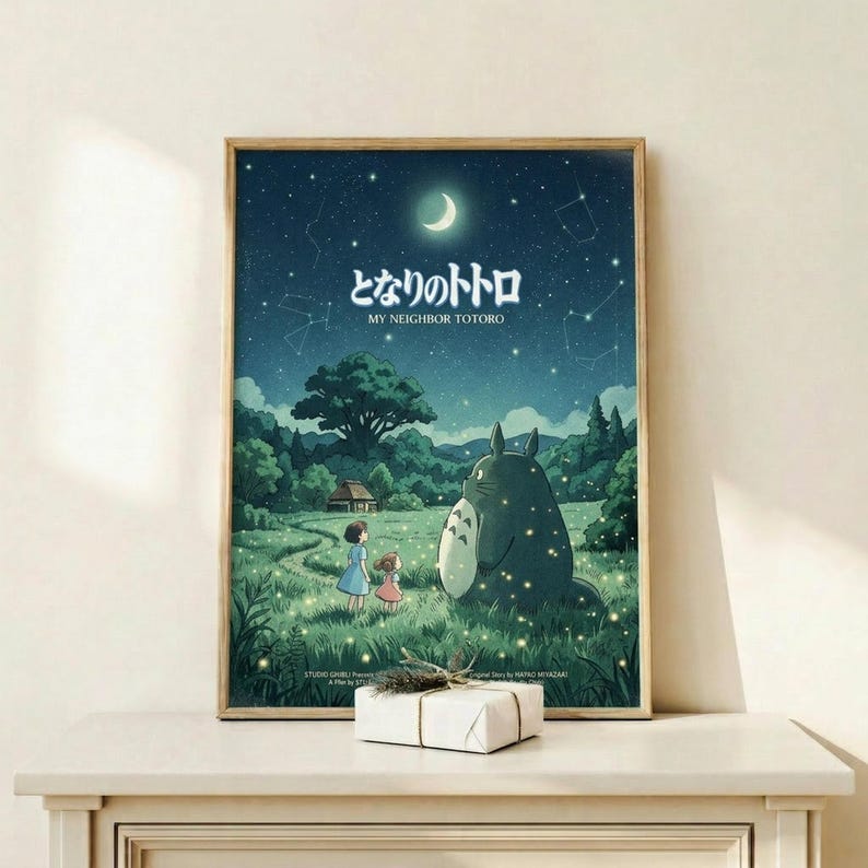 May include: Framed Studio Ghibli My Neighbor Totoro movie poster displayed on light wood shelf with small wrapped gift box. Poster shows nighttime scene with crescent moon, stars, constellations, large gray Totoro character with two children in grassy meadow surrounded by lush green trees. Japanese text at top with English translation below. Artwork features deep blues and greens in whimsical animation style with glowing fireflies scattered throughout scene.