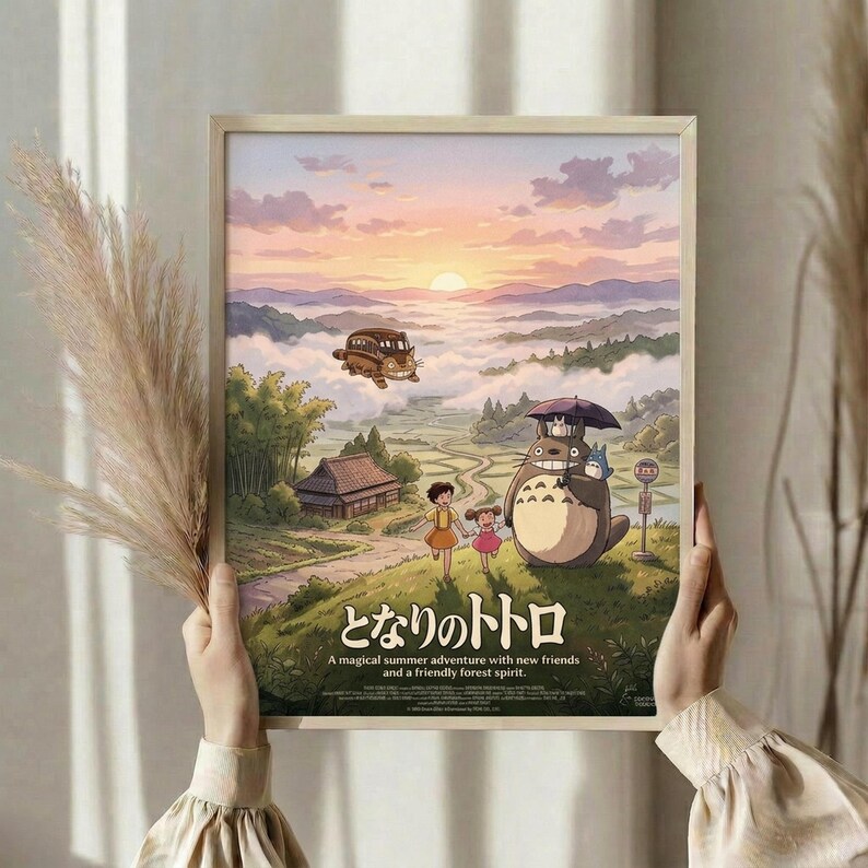 May include: Hands holding framed Studio Ghibli poster in light wood frame against cream curtained window. Artwork shows Totoro with umbrella and children in pastoral landscape with flying bus, mountains, and sunset sky. Japanese text at bottom with English subtitle about magical summer adventure. Dried pampas grass visible on left side.