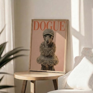 May include: Framed poster featuring a whippet dog wearing a tweed flat cap and fuzzy brown scarf against a pink background, with "DOGUE" text in orange letters at top, displayed on a light wood round side table next to a beige upholstered chair with white throw pillow, green plant leaves visible in corner, soft natural lighting creating shadows on white wall