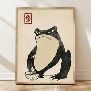 May include: A framed art print depicting a black and white frog seated with a bowl. The frog has a stern expression. A red Japanese character is in the upper left corner. The background is a light beige colour.