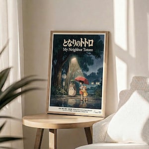 May include: Framed My Neighbor Totoro movie poster in light wood frame, displayed on round wooden side table next to beige upholstered chair. Poster shows iconic rainy bus stop scene with Totoro and children under red umbrella, featuring Japanese text and dark forest atmosphere. Soft natural lighting creates shadows across neutral-toned room with plant visible in corner.