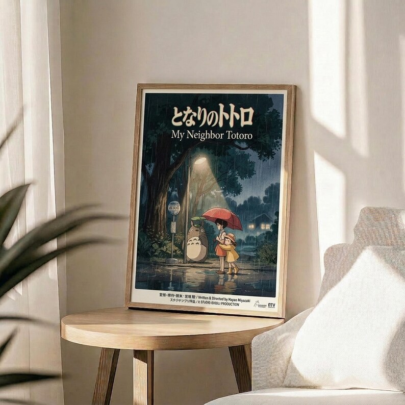 May include: Framed My Neighbor Totoro movie poster in light wood frame, displayed on round wooden side table next to beige upholstered chair. Poster shows iconic rainy bus stop scene with Totoro and children under red umbrella, featuring Japanese text and dark forest atmosphere. Soft natural lighting creates shadows across neutral-toned room with plant visible in corner.