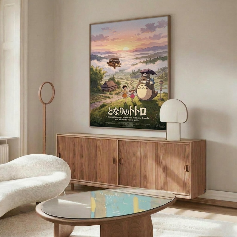 May include: Framed Studio Ghibli My Neighbor Totoro movie poster displayed on white wall above wooden credenza with sliding doors. Poster shows iconic characters including Totoro with umbrella, children, and rural Japanese landscape at sunset with floating airship. Modern living room setting features cream upholstered chair, glass-top oval coffee table, white table lamp, and wooden floor lamp with natural lighting from window.