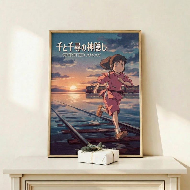May include: Framed Spirited Away movie poster showing animated girl with brown hair in pink outfit running across flooded train tracks at sunset, displayed on light wood shelf next to small white wrapped gift with greenery, against cream wall in minimalist room setting