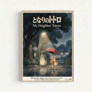Framed poster featuring animated characters from Studio Ghibli's My Neighbor Totoro standing at a rainy bus stop. Two children with a red umbrella wait beside the large gray forest spirit Totoro under a glowing street lamp. The scene shows a nighttime forest setting with heavy rain, puddles reflecting light, and a bus stop sign. Japanese and English text appears at the top, with production credits at the bottom. The poster has a warm wooden frame against a white wall.