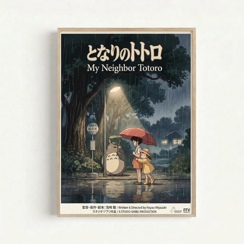 Framed poster featuring animated characters from Studio Ghibli's My Neighbor Totoro standing at a rainy bus stop. Two children with a red umbrella wait beside the large gray forest spirit Totoro under a glowing street lamp. The scene shows a nighttime forest setting with heavy rain, puddles reflecting light, and a bus stop sign. Japanese and English text appears at the top, with production credits at the bottom. The poster has a warm wooden frame against a white wall.