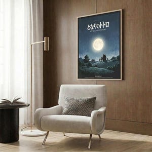 May include: Framed poster on wood-paneled wall showing moonlit night scene with silhouettes under full moon, displayed above modern cream armchair with geometric pillow, brass floor lamp, and dark round side table with open book in contemporary living room setting.
