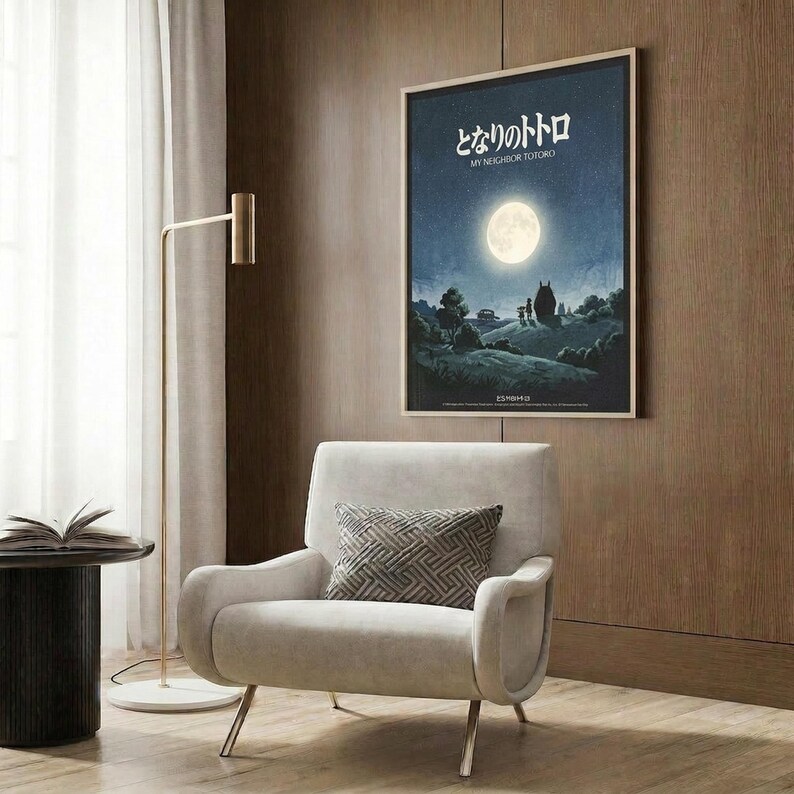 May include: Framed poster on wood-paneled wall showing moonlit night scene with silhouettes under full moon, displayed above modern cream armchair with geometric pillow, brass floor lamp, and dark round side table with open book in contemporary living room setting.