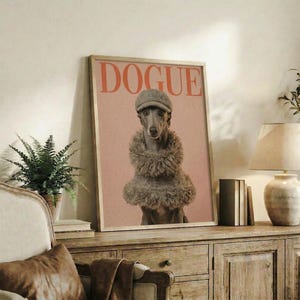 May include: Framed poster featuring a whippet dog wearing a gray knit beanie and fuzzy brown sweater on pink background with "DOGUE" text in red letters, displayed on rustic wooden console table alongside books, ceramic vases, fern plant, and cream upholstered chair in cozy living room setting with white walls.