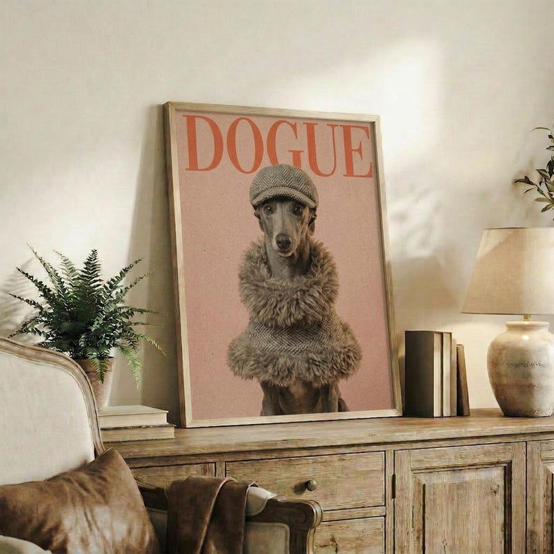 May include: Framed poster featuring a whippet dog wearing a gray knit beanie and fuzzy brown sweater on pink background with "DOGUE" text in red letters, displayed on rustic wooden console table alongside books, ceramic vases, fern plant, and cream upholstered chair in cozy living room setting with white walls.