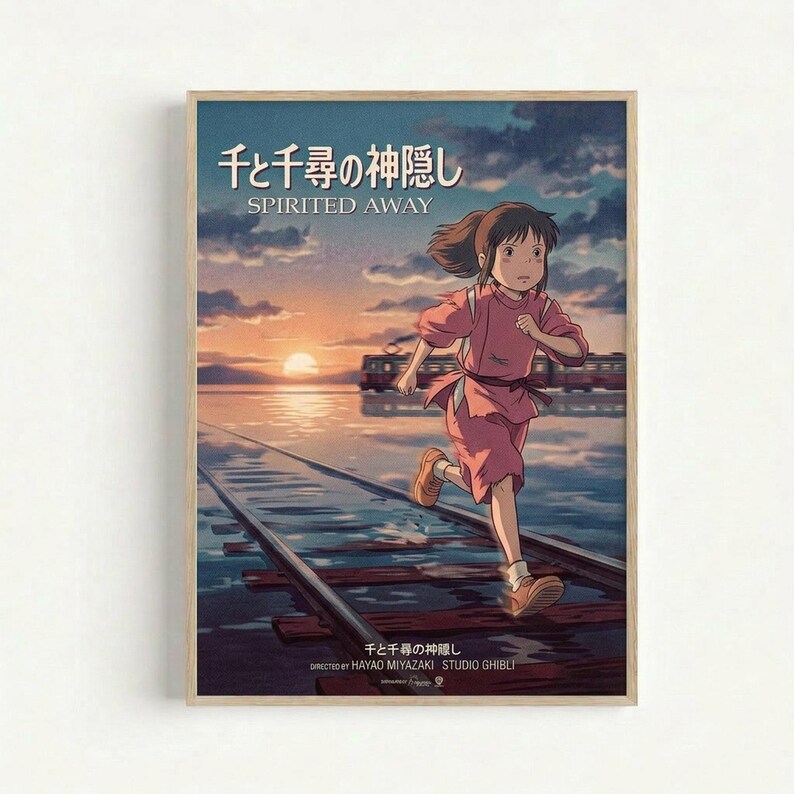 Framed movie poster featuring an animated girl with brown hair running along flooded railway tracks. She wears a pink outfit and sneakers, with a train visible in the background. The scene shows a golden sunset over water with cloudy skies. Japanese text appears at the top with "SPIRITED Away" below it, and "DIRECTED BY HAYAO MIYAZAKI STUDIO GHIBLI" at the bottom. The poster has a light wooden frame and is displayed against a white wall.