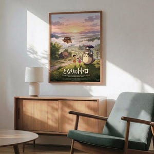 May include: Framed Studio Ghibli Totoro movie poster hanging on white wall above wooden sideboard. Poster shows iconic gray forest spirit character with children on rural path, flying bus in cloudy mountain landscape at sunset. Mid-century modern room with teal upholstered chair, round wood coffee table, white ceramic lamp. Natural sunlight creates geometric shadows on wall.