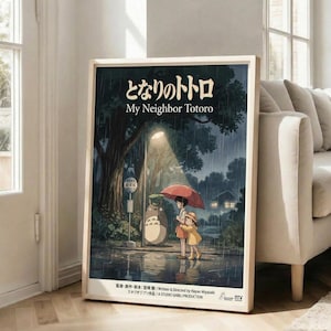May include: Framed My Neighbor Totoro movie poster leaning against white wall beside beige sofa in bright living room with wooden floors. Poster shows iconic rainy bus stop scene with Totoro, Mei, and Satsuki under red umbrella beneath glowing streetlight, surrounded by dark trees and atmospheric lighting in Studio Ghibli's signature animation style.