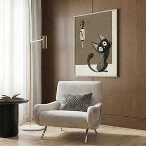 May include: Modern living room scene with beige armchair and geometric pillow, gold floor lamp, dark round side table with open book, and wood-paneled wall. Framed artwork shows black cat character with large white eyes sitting upright, accompanied by Japanese characters and decorative lantern illustration on neutral beige background.