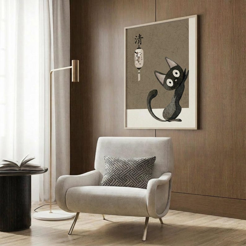 May include: Modern living room scene with beige armchair and geometric pillow, gold floor lamp, dark round side table with open book, and wood-paneled wall. Framed artwork shows black cat character with large white eyes sitting upright, accompanied by Japanese characters and decorative lantern illustration on neutral beige background.