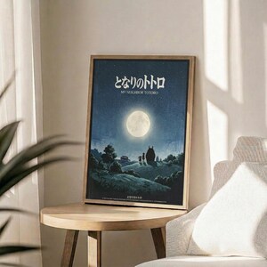 May include: Framed movie poster with Japanese text and English "My Neighbor Totoro" title displayed on wooden round side table next to beige upholstered chair. Poster features nighttime scene with full moon, starry blue sky, and silhouettes including large Totoro character and two children on hillside landscape. Natural lighting creates soft shadows across neutral-toned room setting with plant visible in corner.