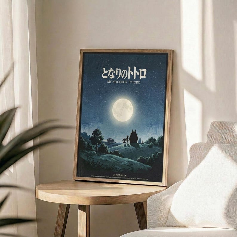 May include: Framed movie poster with Japanese text and English "My Neighbor Totoro" title displayed on wooden round side table next to beige upholstered chair. Poster features nighttime scene with full moon, starry blue sky, and silhouettes including large Totoro character and two children on hillside landscape. Natural lighting creates soft shadows across neutral-toned room setting with plant visible in corner.