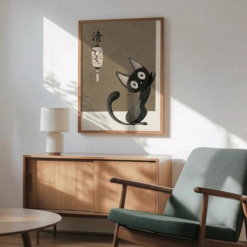 May include: Framed poster featuring a black cat character with wide eyes sitting beneath a white paper lantern with Japanese text, displayed in a mid-century modern living room with sage green upholstered chair, wood sideboard, white ceramic lamp, and natural sunlight casting shadows on white walls.