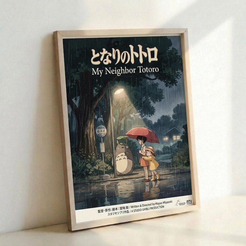 May include: Framed poster featuring My Neighbor Totoro movie artwork displayed against a white wall. The poster shows children with umbrellas meeting Totoro at a rainy bus stop under large trees, rendered in Studio Ghibli's distinctive animated style with deep blues and greens. Japanese text appears at the top with English title below. Natural wood frame surrounds the artwork.