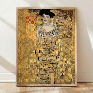 May include: A framed print of a Gustav Klimt painting. The artwork features a woman in a detailed, golden dress with geometric patterns and a decorative collar. The background is filled with gold leaf and intricate designs, creating a rich, textured appearance.