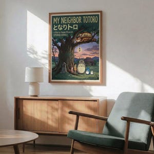 May include: Studio Ghibli My Neighbor Totoro movie poster displayed in modern living room with mid-century wooden credenza, sage green upholstered chair, and ceramic table lamp. Poster shows iconic Totoro characters under large tree with Catbus above, rendered in warm earth tones with green and purple sky. Natural sunlight streams across white walls creating geometric shadows.