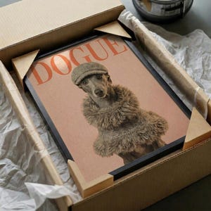 May include: Framed poster featuring a whippet dog wearing a flat cap and fur coat on pink background with "DOGUE" text in coral letters, displayed in black frame inside shipping box with protective packaging materials on white surface