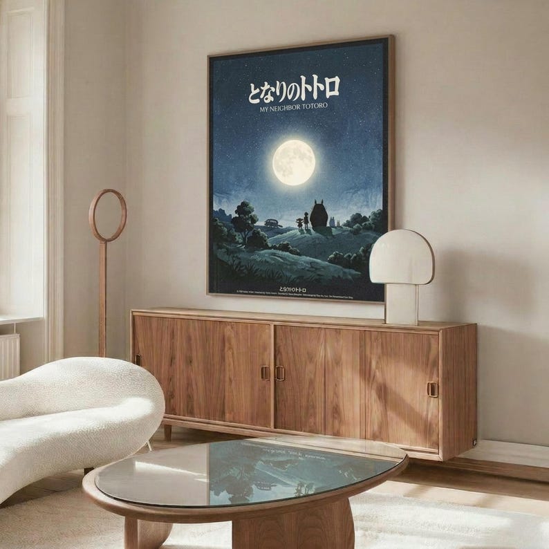 May include: Framed My Neighbor Totoro movie poster displayed above a wooden sideboard in a minimalist living room. The poster features a nighttime scene with a full moon, starry sky, and silhouetted Totoro character with two children on a hill. Modern white curved chair and glass-top coffee table in foreground. Warm wood tones throughout furniture, cream walls, and soft natural lighting create a cozy mid-century modern aesthetic.