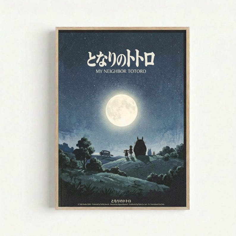 A framed poster featuring a moonlit night scene with two small children standing beside the large, round silhouette of Totoro on a hillside. The full moon illuminates a starry blue sky above rolling countryside with trees, and a small bus is visible in the distance. Japanese text and "MY NEIGHBOR TOTORO" appear at the top in white lettering. The artwork has a dreamy, nostalgic animation style in blue and cream tones, displayed in a light wood frame against a white wall.