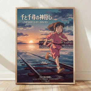 May include: Framed movie poster leaning against white wall on wooden floor. Features animated girl with brown hair in pink outfit running on flooded train tracks toward sunset. Train visible in background across water. Japanese and English text at top reads "Spirited Away." Directed by Hayao Miyazaki and Studio Ghibli credits at bottom. Natural wood frame, soft lighting creates shadows on wall.