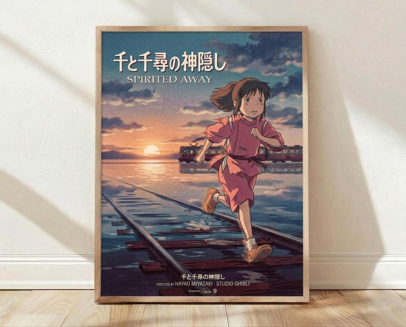 May include: Framed movie poster leaning against white wall on wooden floor. Features animated girl with brown hair in pink outfit running on flooded train tracks toward sunset. Train visible in background across water. Japanese and English text at top reads "Spirited Away." Directed by Hayao Miyazaki and Studio Ghibli credits at bottom. Natural wood frame, soft lighting creates shadows on wall.