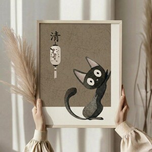May include: Person in cream sweater holding framed artwork featuring a stylized black cat with large white eyes sitting upright, alongside Japanese characters and a decorative white lantern with floral design. The artwork has a textured beige background in a light wood frame. Dried wheat stalks visible on the left side against soft neutral curtains and window lighting.