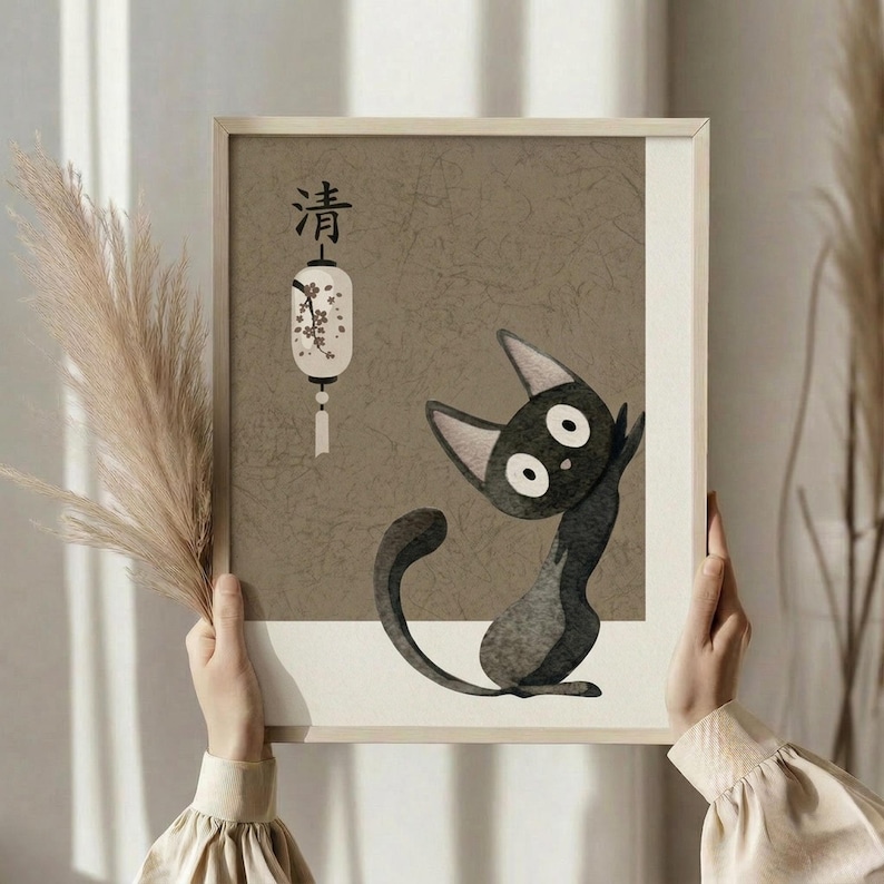 May include: Person in cream sweater holding framed artwork featuring a stylized black cat with large white eyes sitting upright, alongside Japanese characters and a decorative white lantern with floral design. The artwork has a textured beige background in a light wood frame. Dried wheat stalks visible on the left side against soft neutral curtains and window lighting.