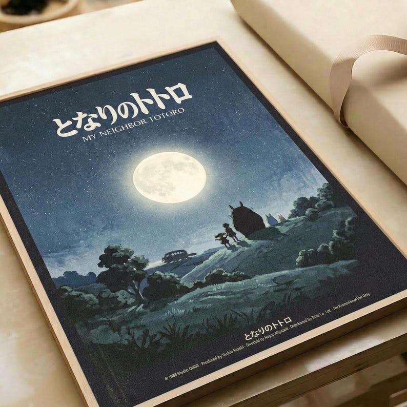 My Neighbor Totoro movie poster featuring a nighttime landscape scene with a large glowing full moon in a starry blue sky. Silhouettes of Totoro, a small child, and a Catbus are positioned on rolling green hills dotted with trees. Japanese text appears at the top with English subtitle below. The poster has a nostalgic, dreamlike quality with deep blues and moonlit tones, displayed flat against cream-colored wrapping paper with a beige ribbon visible at the edge.