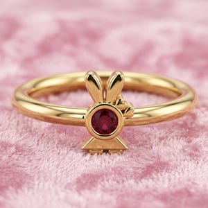 Könnte beinhalten: Ein goldfarbener Ring mit einem Hasenmotiv und einem runden, roten Edelstein. Der Hase hat aufrechte Ohren und ein kleines Herz. Der Ring liegt auf einem rosa Hintergrund.