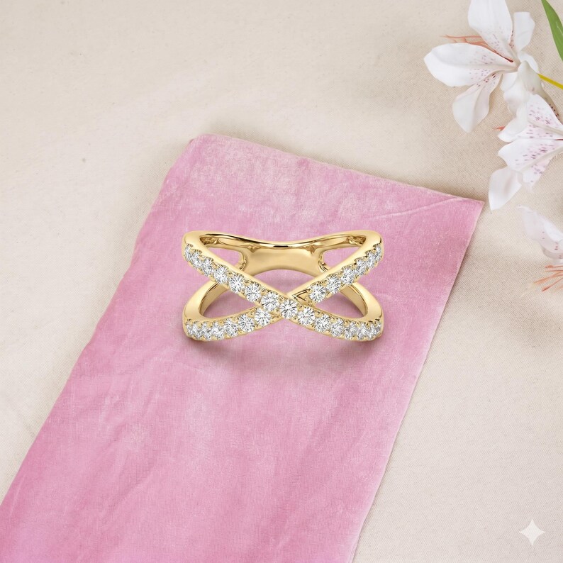 Op de afbeelding: Een goudkleurige crossover ring met een dubbel X-ontwerp. De ring is bezet met kleine, sprankelende witte edelstenen langs de banden. De ring wordt gepresenteerd op een roze ondergrond, met witte bloemen op de achtergrond.