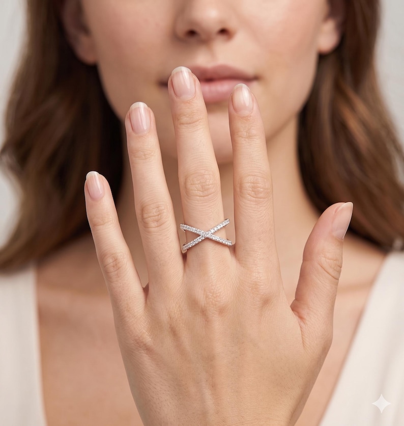 Op de afbeelding: Een zilverkleurige ring met een kruisontwerp, versierd met kleine, sprankelende stenen. De ring wordt getoond op een hand, wat de elegante en moderne stijl benadrukt. Het ontwerp van de ring is een eigentijdse interpretatie van een klassieke look.