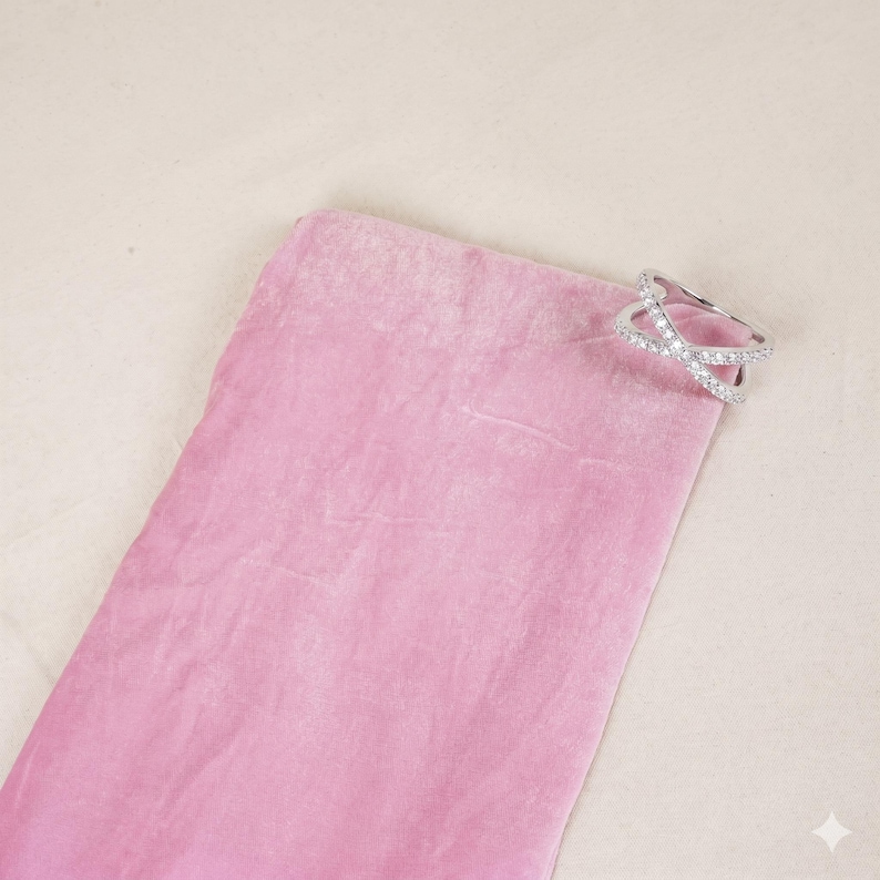 Op de afbeelding: Een zilverkleurige ring met een kruisvormig ontwerp, versierd met kleine, heldere edelstenen. De ring ligt op een roze fluwelen zakje, tegen een neutrale achtergrond. Het ontwerp van de ring bestaat uit twee elkaar kruisende banden, die een X-vorm cre&euml;ren.