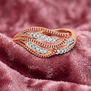 Könnte beinhalten: Ein Ring aus Roségold mit einem geschwungenen, mehrschichtigen Design aus texturierten, gerippten Metallbändern. Der Ring ist mit kleinen, klaren, rund geschliffenen Steinen besetzt, die entlang der wellenförmigen Metallsegmente verlaufen.