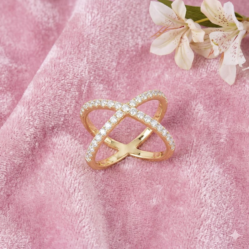 Op de afbeelding: Een goudkleurige ring met een kruisvormig ontwerp, bezet met kleine, heldere edelstenen. De ring ligt op een roze fluwelen achtergrond, met witte en gele bloemen op de achtergrond. Het ontwerp van de ring is een moderne interpretatie van een klassieke stijl.